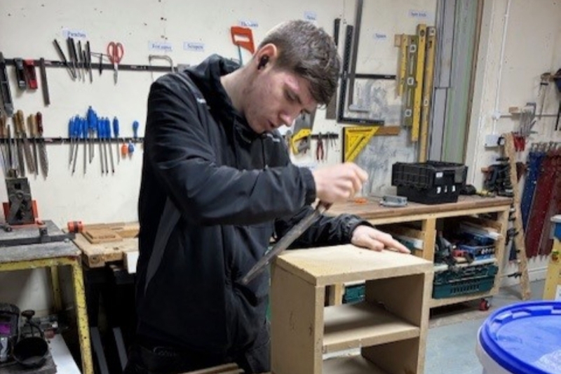 Tate in a workshop using a hand tool to smooth the edges of a small wooden shelving unit. In the background, a wall is organized with various hand tools, including screwdrivers, saws, and levels.