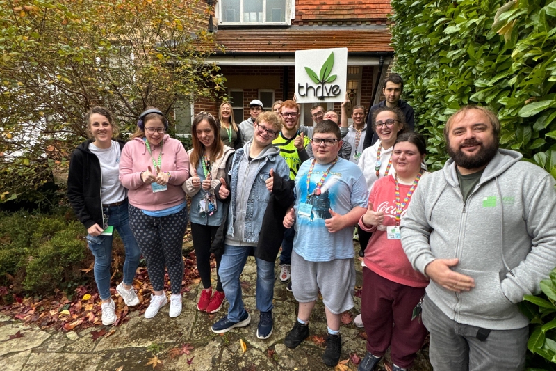 A group of young adults and staff standing outside a brick building, smiling and giving thumbs up. They are holding a sign that reads “Thrive”.